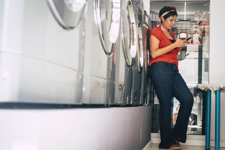 Young woman standing and using mobile phone while wait her clothes washed in a laundromat service shop in the city - modern lifestyle too busy for business career - technology for peopleの写真素材