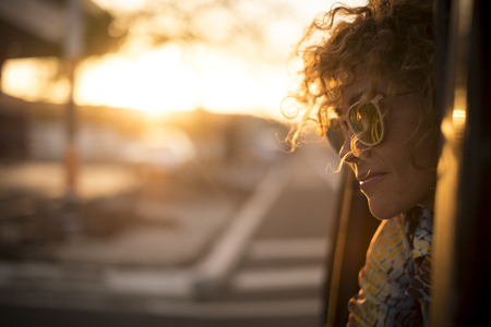 Beautiful sunny sunset time portrait for cute lady smiling and enjoying the feeling with the freedom outdoor - curly hair in the wind and sunglasses for travel concept imageの写真素材