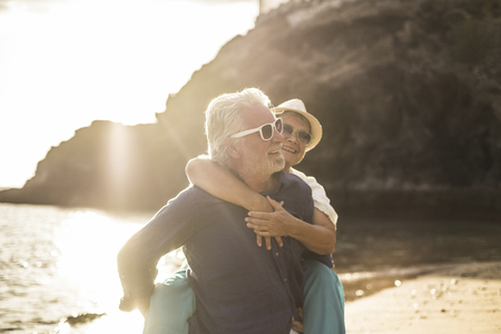 adults caucasian people couple in love with man carrying the woman and both smile and laugh a lot - sunset and backlight at the beach - vacation lifestyle married forever concept for happy matures retiredの写真素材