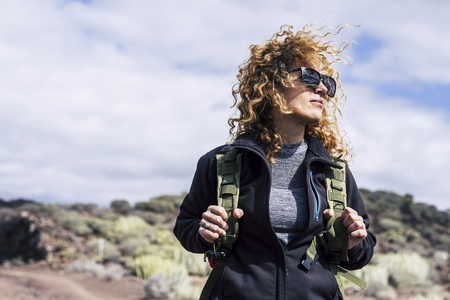 Beautiful curly hair middle age caucasian woman with backpack in trekking activity outdoor - female hiker at the mountain looking - blue sky background and alternative vacation conceptの写真素材