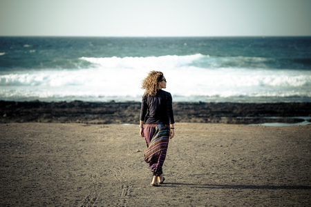 One woman walking at the beach to the ocean and waves enjoying the feeling of freedom and nature with wind in the curly hair - people in outdoor vacational activityの写真素材