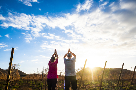 Yoga and meditation position with two aged senior man and woman together looking and giving thanks to the sun in early morning or golden sunset - outdoor healthy activity for nice lifestyle peopleの写真素材