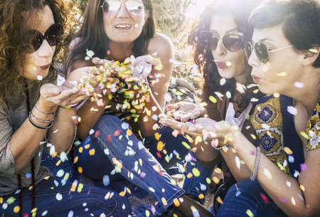 Group of attractive middle age young women playing and blowing out a lot of coloured confetti in the air - celebrate and having fun together with friendship - party outdoor concept with sun backlightの写真素材