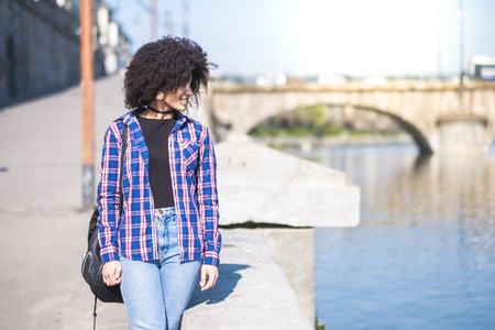 Hipster beautiful cheerful young black girl walking inthe city enjoying the outdoor leisure activity near a river - student with backpack enjoy the spring - african ethnich curly hair - young people happyの写真素材