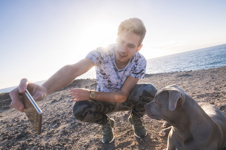 Best friends together young caucasian man and grey beautiful amstal dog taking selfie picture with smart phone outdoor with sunlight sunset in background - ocean and vacation conceptの写真素材