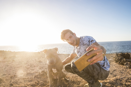 Happy couple of alternative millennial friends man and dog taking selfie picture with modern phone during a sunset in outdoor leisure activity together neat the ocean - sunny backlight and pet therapy lifestyleの写真素材