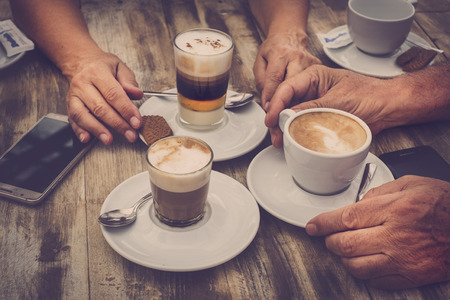 Close up aged caucasian hands with coffee cappuccino at the bar for breakfast - wooden table and vintage romantic color tones - mobile phone with the people - love and couple conceptの写真素材