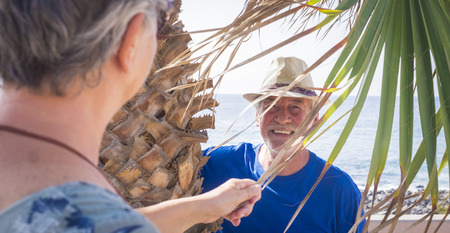 close-up of a 70-year-old man laughing under a palm tree playing hide-and-seek. Vacation, fun concept - Cheerful caucasian aged people in outdoor leisure activity togetherの写真素材