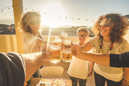 Group of friends clinking all together having fun in friendship outdoor on the rooftop terrace with city and sea view during summer dinner at the sunset at home - vacation and enjoying lifestyleの写真素材
