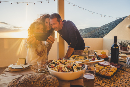 Group of friends caucasian people celebrate together outdoor in the terrace with a table full of food and drinks and take selfie having fun - friendship concept for adult man and women enjoying the meals and celebrate the summer timeの写真素材