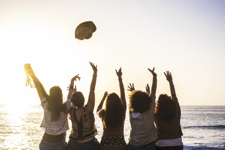 Summer and happiness for travel and freindship concept with group of happy girls viewed from rear enjoying and celebrating the sunset and the sea together launching hat and giving upの写真素材