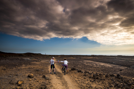 Couple middle age and senior ride with mountain bike ona path in the desert. Beautiful outdoor in background with colors and dramatic clouldy sky at the sunset - leisure sport activity conceptの写真素材