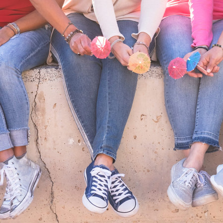 Close up unrecognizable group of women legs with young sneakers shoes and little cocktail umbrella for summer friendship concept together sitting on a wall - casual and coloured clothesの写真素材