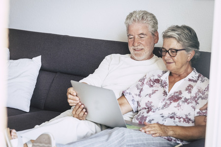 Beautiful caucasian aged senior couple at home watching movie or searching on the web sitting on the sofa with a laptop on the legs - indoor home technology concept for old peopleの写真素材