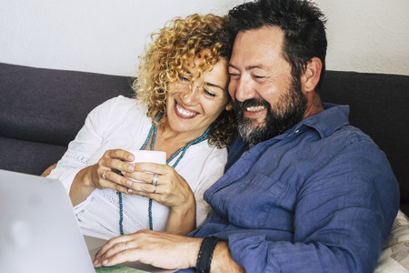 Cheerful beautiful happy caucasian couple middle age at home sitting on the couch using a technology device laptop  - internet and leisure activity indoor for people smiling and enjoying the timeの写真素材