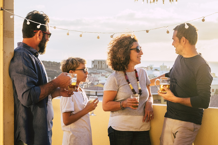 Group of middle age people caucasian men and woman drink together on the terrace with city view having fun and friendship - blonde kid with them - outdoor home leisure activity for friendsの写真素材