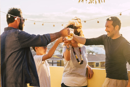Group of friends toasting and clinking glasses with beer and orange juice together during the sunset on the roof terrace with city view - mixed ages and generations having fun celebratingの写真素材