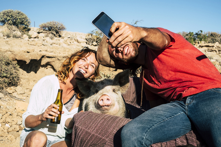 Alternative family or friends with young man and woman smiling while take a selfie with a domestic pig in the middle - everybody smile for the camera - sunny day and unusual use of phone camera technology - funny animalの写真素材
