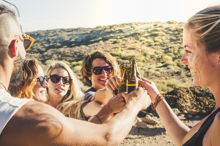 Group of young caucasian people boys and girls clinking together beer bottles in natural celebration day - enjoying outdoor and friendship lifestyle - happiness and joyful for youth men and womenの写真素材