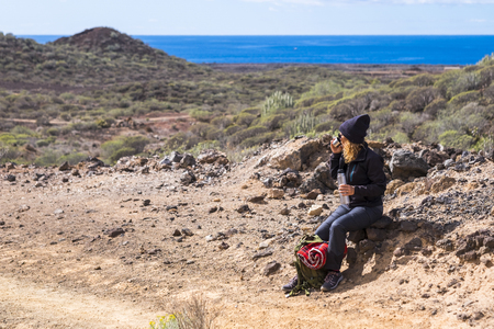 Curly cute woman doing trekking and hking in rock mountains with ocean in background - resting with drink - backpacker lifestyle for alternative leisure activity - people in outdoorの写真素材