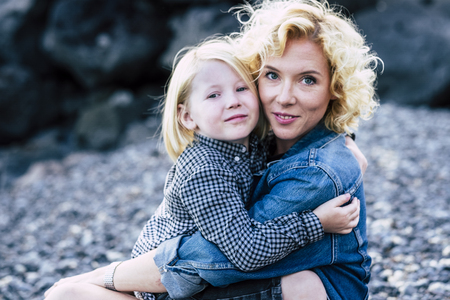Beautiful caucasian young blonde mother hug her little son with long hair with love and affection - family outdoor - cheerful mom and children have fun together - defocused bokeh backgroundの写真素材