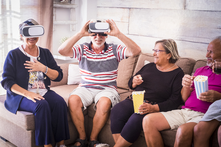 Group of aged old friends have fun at home with vr goggled headset and pop corn - party celebrating time with laughs and friendship for caucasian mature peopleの写真素材