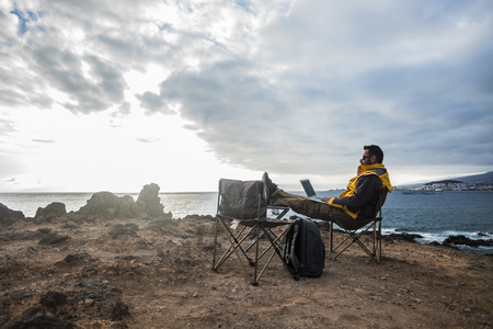 Digital noman and traveler middle age adult man sitting and working with internet and laptop in totally fredom in front of the ocean - backpacker lifestyle and alternative office for happy lifeの写真素材