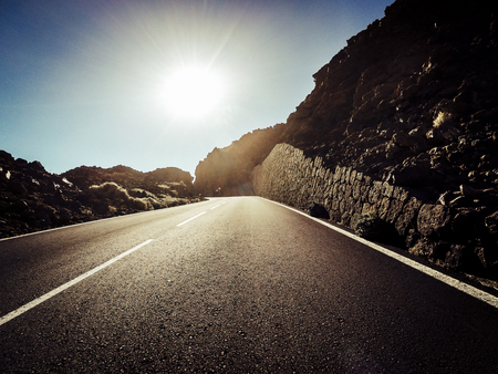 Long way road at the mountain with sun in front and sunlight effect - ground point of view with black asphalt and white lines - driving and travel conceptの写真素材
