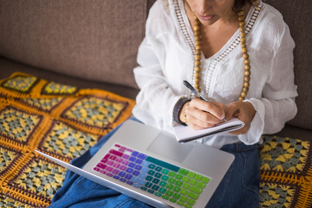 Technology using a computer laptop at home concept with concentrated young caucasian woman at work on the sofa in leisure indoor activity - Writing notes on paper - patchwork handmade cover coloursの写真素材