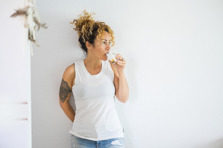 Attractive curly blonde middle age cute woman drinking water and lemon at home with white wall in background - healthy lifestyle for cacuasian people - modern tatoo girlの写真素材
