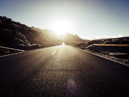 Long way road at the mountain with sun in front and sunlight effect - ground point of view with black asphalt and white lines - driving and travel conceptの写真素材