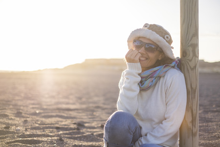 Cheerful beautiful middle age caucasian lady smiling and looking at the camera with golden sunset in background sitting at the beach in outdoor leisure activity or independent vacationの写真素材