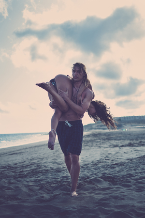 Vintage dark colors for beautiful caucasian couple boy and girl at the beach enjoying and having fun - man carrying woman in summer vacation lifestyle - blue tonesの写真素材