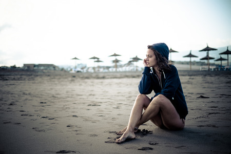 Cute lonely caucasian young girl sitting at the beach on the sand looking the open space and thinking - closed resort with sun umbrelas in background - cold tones and colorsの写真素材