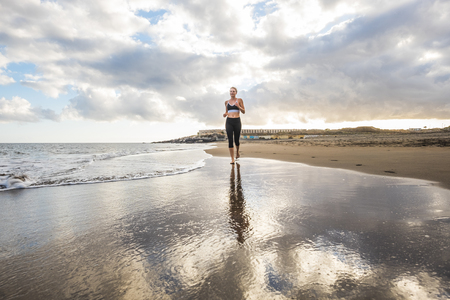 Attractive blonde young woman running at the beach on the shore - scenic place for summer sport fitness activity for people - healthy lifestyle with outdoor workout jogging near the seaの写真素材