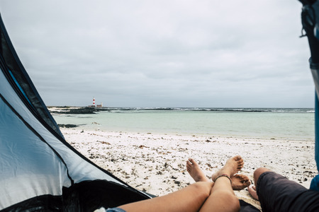 Legs of caucasian couple enjoying the alternative vacation with free camping and tent at the beach with ocean and lighthouse in front - freedom and outdoor leisure activity for alternative travelerの写真素材