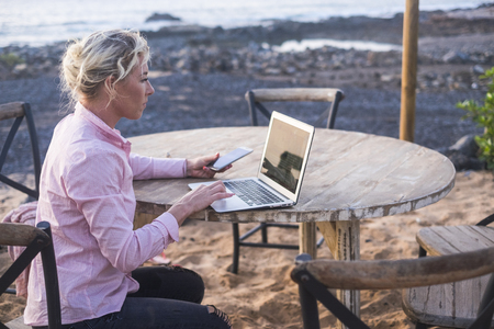 digital nomad concept with blonde free beautiful caucasian middle age woman working at the laptop using cellular phone too sitting on a wood table near the ocean - freelance free to work everywhere with technologyの写真素材