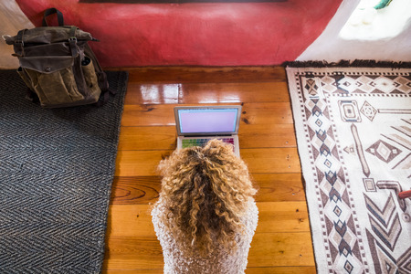 Curly blonde woman working at the laptop lay down on the floor in hotel or home room viewed from above - lady using computer notebook during travel in a coloured houseの写真素材
