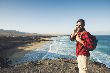 Standing caucasian man calling at the phone on a isolate cliff with ocean waves view - backpacker alternative lifestyle vacation for people love nature and outdoors - technology and adventure conceptの写真素材