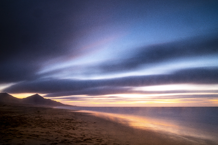 Beautiful landscape in long exposure at the beach during coloured cloudy sunset with mountains in background - wild and scenic vacation concept - cute outdoor nature with colors and ocean horizonの写真素材