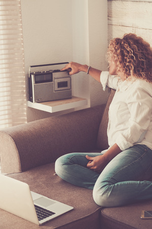 Fashion cute curly young woman at home using old and new technology sitting on the sofa - vintage concept and used radio from '80s - people in the house in indoor leisure activity listening musicの写真素材