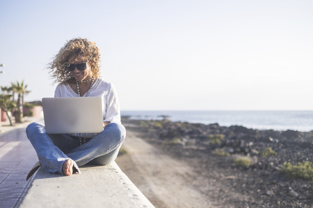Curly attractive young caucasian woman work at the laptop in outdoor near the beach - internet everywhere and freelance digital nomad concept for free people enjoying the lifestyleの写真素材
