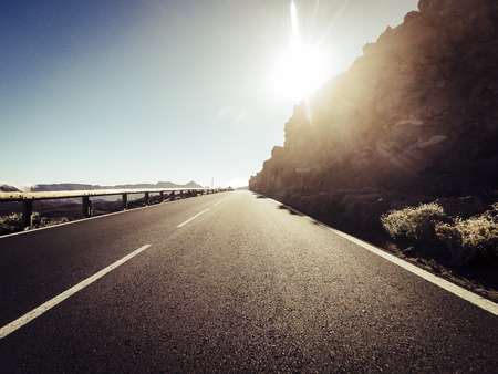 Long way road at the mountain with sun in front and sunlight effect - ground point of view with black asphalt and white lines - driving and travel conceptの写真素材