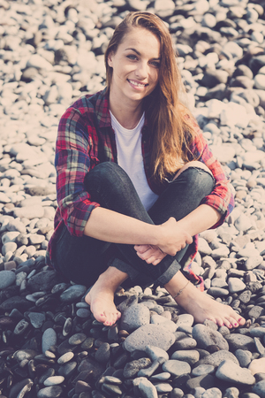 Portrait of young caucasian hipster woman sitting on the rocks in summer sunny day smiling at the camera with happiness - tourist and vacation conceptの写真素材
