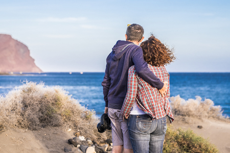 Adult caucasian couple viewed from back hug with love enjoying and looking the ocean and the coast during vacation - tourist and tourism concept with middle age people together in relationshipの写真素材