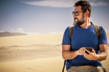 Senior caucasian nice man with beaard hipster style use cellular phone with internet connection in the desert dunes alone smiling and enjoying the wild outdoor place - alternative travel lifestyle conceptの写真素材