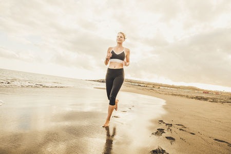 Beautiful blonde yong woman run at the beach to train and follow a healthy active lifestyle - enjoying running outdoor on the shore in barefoot way - smile girl doing joggingの写真素材