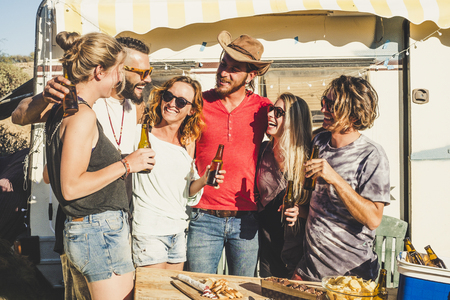 Group of cheerful people youthful young men and women have fun together in friendship with beers - old vintage alternative vacation caravan in background - rural place and guys laughingの写真素材