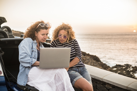Couple of people women friends use together a laptop computer sitting on the back of convertible car and sunset on the ocean in background - work everywhere and travel vacation plan conceptの写真素材