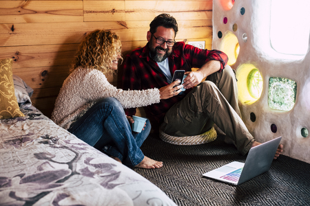 Adult couple enjoying technology at home or hotel traveling with looking a mobile phone device screen and using a laptop internet connected on the floor - alternative people conceptの写真素材
